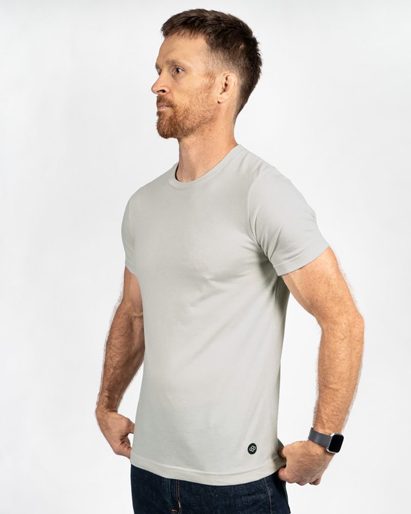 A man poses in profile wearing a light gray short-sleeve t-shirt and dark jeans showcasing a minimalist style and casual appearance