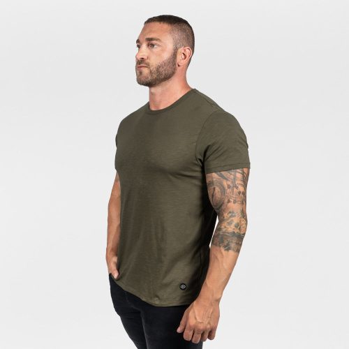 A man with a defined physique stands against a plain white background, wearing an olive green crew neck t-shirt and black pants, creating a minimalist aesthetic.