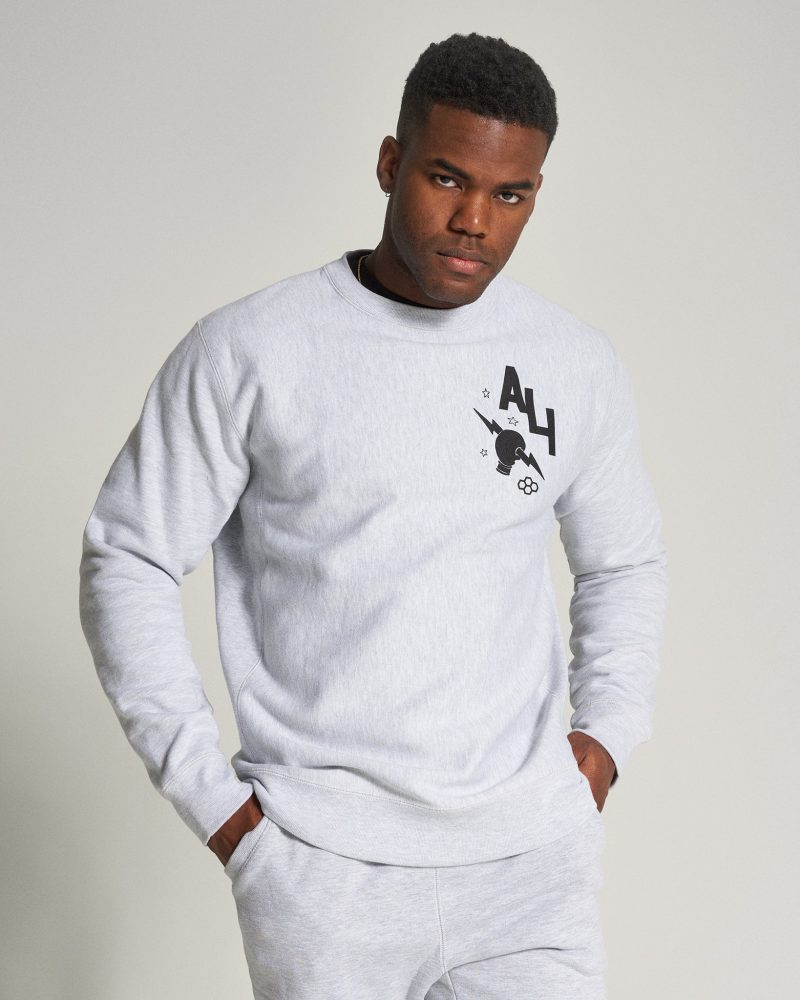 A young man poses confidently in a light gray sweatshirt with a graphic print complemented by matching sweatpants against a neutral background