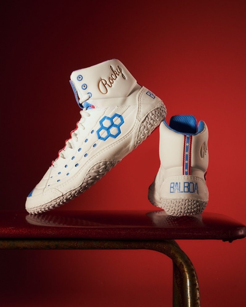 A pair of white high-top sneakers featuring blue accents gold embroidery and a distinctive tread design presented against a bold red background