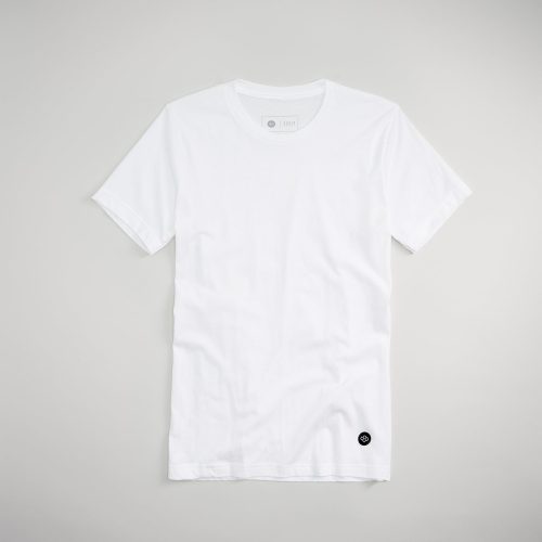 A plain white t-shirt displayed flat against a muted gray background featuring a subtle logo on the bottom left