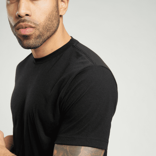 A close-up portrait of a man with a beard and tattoos wearing a fitted black t-shirt against a neutral background